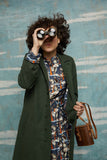 A person with curly hair wearing a Palava Trenchcoat in dark green from BUTIK KUBIK looks through binoculars, holding a brown bag. They are against a blue and white painted wall, sporting a colorful dress underneath.