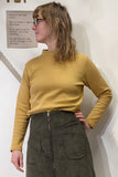 A person with shoulder-length hair and glasses gazes sideways, wearing a mustard yellow Kubik sweater waffle by BUTIK KUBIK and an olive green pocketed skirt. Made from organic fabric, their outfit radiates 60s charm as they stand against a white wall with partially visible text beside them.
