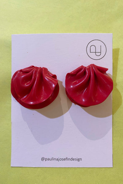 A pair of red, dumpling-shaped Paulina Josefin design earrings by BUTIK KUBIK in polymer clay, shown on a white card with logo and @paulinajosefindesign, set against a light yellow background.