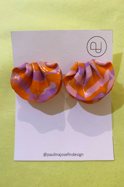A pair of crinkled, fan-shaped Paulina Josefin design earrings by BUTIK KUBIK in orange and lavender patterns are displayed on a white card with the Paulina Josefin logo and Instagram handle @paulinajosefindesign, against a light yellow background.