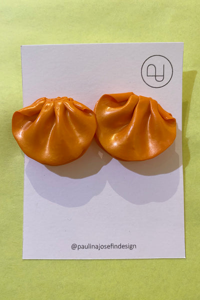 A pair of shiny orange Paulina Josefin design earrings by BUTIK KUBIK, resembling dumplings, shown on a branded card with @paulinajosefindesign and logo, set against a light yellow background.
