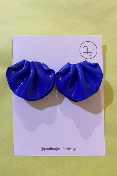 A pair of bright blue, ruffled Paulina Josefin design earrings by BUTIK KUBIK are displayed on a white card with the Paulina Josefin logo and @paulinajosefindesign, set against a light yellow background.