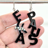 En hand håller Favoretro Earrings Fikapaus Plain Black från BUTIK KUBIK – två laserskurna svarta akrylörhängen formade som bokstäver: “FA” på ena och “PAS” på andra, med vertikalt staplade bokstäver.