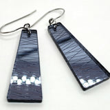 A pair of Favoretro earrings sketch by BUTIK KUBIK, featuring trapezoid-shaped plexiglass in a glossy thundercloud gray with crisscrossing etched lines, attached to surgical steel hooks and displayed on a white background.
