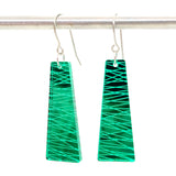 Two green trapezoid-shaped plexiglas Favoretro earrings sketch from BUTIK KUBIK with silver hooks hang from a metal rod. The earrings feature a shiny surface with intersecting diagonal lines forming a geometric pattern, and the hooks are made of kirurgiskt stål for comfortable, hypoallergenic wear.