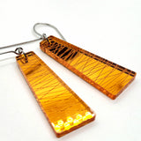 Two rectangular, amber-colored plexiglass “Favoretro earrings sketch” by BUTIK KUBIK, featuring subtle diagonal line patterns and surgical steel hooks, are displayed on a white background.