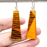 A hand holds two Favoretro earrings sketch by BUTIK KUBIK—orange trapezoidal plexiglass earrings with etched diagonal lines, hanging from surgical steel hooks on a metal bar.