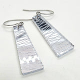 A pair of Favoretro earrings sketch by BUTIK KUBIK, featuring silver-tone, trapezoid-shaped dangles crafted from surgical steel with etched diagonal and horizontal lines, displayed on a white background.