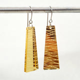 Two trapezoid-shaped Favoretro earrings sketch by BUTIK KUBIK, crafted from plexiglas in yellow and amber with geometric line patterns, hang from a rod of kirurgiskt stål against a plain white background.
