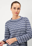 A person with brown hair in a bun stands against a plain backdrop wearing the Jumperfabriken Saralisa Jumper Navy by BUTIK KUBIK. The cotton top has a blue and white pattern, long sleeves, and a round neckline.