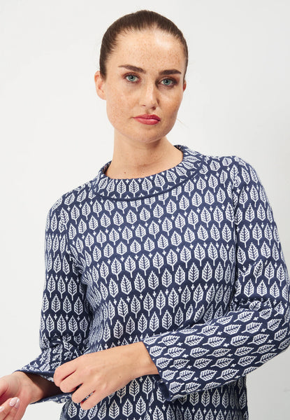 A person with brown hair in a bun stands against a plain backdrop wearing the Jumperfabriken Saralisa Jumper Navy by BUTIK KUBIK. The cotton top has a blue and white pattern, long sleeves, and a round neckline.