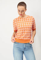 A person wearing the BUTIK KUBIK Jumperfabriken Bess Jumper in orange, featuring a floral pattern and short sleeves, pairs it with light blue jeans while standing against a plain background, looking down and adjusting the hem.