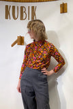 A blond person in glasses wears the BUTIK KUBIK Kubik sweater retro 1974 and gray pants, standing before a white wall with "KUBIK" above and two small wooden shelves mounted on it.