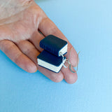 A hand holds a pair of Becca design Bok earrings by BUTIK KUBIK, shaped like small blue books with white pages, resting on a light blue textured surface.