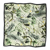 The BUTIK KUBIK Sneaky fox silk scarf Fern features a green floral print with leaves, ferns, white flowers, and yellow birds on a light background, finished with a slim black border.