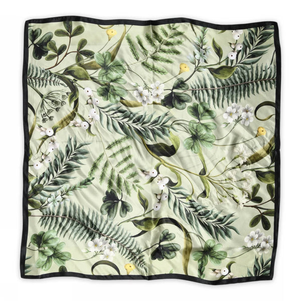 The BUTIK KUBIK Sneaky fox silk scarf Fern features a green floral print with leaves, ferns, white flowers, and yellow birds on a light background, finished with a slim black border.
