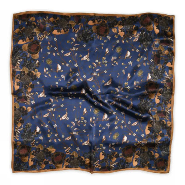 The Sneaky fox silk scarf Kayla by BUTIK KUBIK features a deep blue background with birds, flowers, leaves, and butterflies in earthy tones, framed by a brown border. Made from 100% silk for a luxurious feel.
