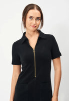 A woman with fair skin and brown hair tied back wears the short-sleeved Jumperfabriken Lovisa black dress by BUTIK KUBIK. Made from 100% cotton, it features a gold zipper, two front pockets, and effortless style for beachwear comfort.