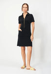 A woman stands against a plain background in the Jumperfabriken Lovisa black dress by BUTIK KUBIK—100% cotton terry, front zipper, pockets—perfect for summer. She faces forward with hands in pockets, wearing yellow slide sandals.
