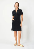 A woman stands against a plain background in the Jumperfabriken Lovisa black dress by BUTIK KUBIK—100% cotton terry, front zipper, pockets—perfect for summer. She faces forward with hands in pockets, wearing yellow slide sandals.