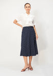 A woman stands against a plain background wearing a white short-sleeve blouse with a bow at the neck, and the BUTIK KUBIK Jumperfabriken Sunvi Dot Skirt—a navy polka dot viscose skirt with an elastic waist—paired with brown heeled sandals.