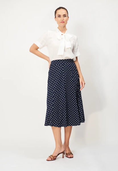 A woman stands against a plain background wearing a white short-sleeve blouse with a bow at the neck, and the BUTIK KUBIK Jumperfabriken Sunvi Dot Skirt—a navy polka dot viscose skirt with an elastic waist—paired with brown heeled sandals.
