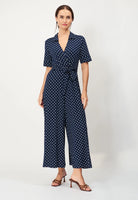 A person models the BUTIK KUBIK Jumperfabriken Blake Dot Jumpsuit in Midnight Blue, featuring short sleeves and a belted waist, paired with brown heeled sandals, against a plain white background.