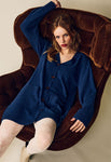 A woman with light skin and red hair, wearing the Jumperfabriken cardigan Asta by BUTIK KUBIK and white patterned tights, sits relaxed on a plush brown chair, looking directly at the camera with a neutral expression.