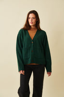 A woman with long brown hair stands against a beige background, wearing the BUTIK KUBIK Jumperfabriken cardigan Estrid over a tan ribbed turtleneck and black pants. She is facing forward with a relaxed expression.