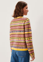 A woman with shoulder-length brown hair, seen from behind, wears the BUTIK KUBIK Jumperfabriken cardigan AnnaLisa with retro yellow, red, blue, and green stripes and blue jeans, standing against a plain white background.