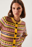 A woman with brown hair models the Jumperfabriken cardigan AnnaLisa from BUTIK KUBIK, a merino wool sweater with a colorful retro pattern, yellow trim, and large brown buttons, standing against a plain white background.