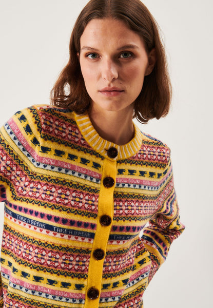 A woman with brown hair models the Jumperfabriken cardigan AnnaLisa from BUTIK KUBIK, a merino wool sweater with a colorful retro pattern, yellow trim, and large brown buttons, standing against a plain white background.