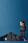 A woman with long brown hair sits on the floor against a blue background, wearing the Jumperfabriken Wednesday Dress from BUTIK KUBIK, black knee-high boots, and blue earrings. She gazes calmly to the right.