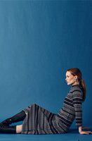 A woman with long brown hair sits on the floor against a blue background, wearing the Jumperfabriken Wednesday Dress from BUTIK KUBIK, black knee-high boots, and blue earrings. She gazes calmly to the right.