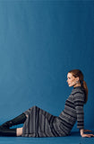 A woman with long brown hair sits on the floor against a blue background, wearing the Jumperfabriken Wednesday Dress from BUTIK KUBIK, black knee-high boots, and blue earrings. She gazes calmly to the right.