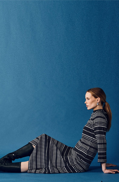 A woman with long brown hair sits on the floor against a blue background, wearing the Jumperfabriken Wednesday Dress from BUTIK KUBIK, black knee-high boots, and blue earrings. She gazes calmly to the right.