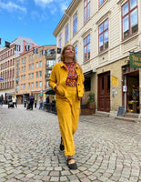 A woman in the BUTIK KUBIK Kubik Overshirt mustard and a patterned top walks confidently down a cobblestone street lined with old brick and wooden buildings on a bright day.