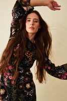 A woman with long brown hair models the BUTIK KUBIK Jumperfabriken dress Lenita Black, striking a confident pose with one arm raised and looking at the camera against a neutral background.
