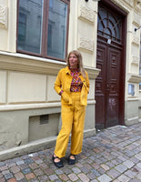 A woman stands on a cobblestone street in front of a tan building, wearing BUTIK KUBIKs Kubik corduroy trousers in bright yellow, styled as a pantsuit with a colorful patterned top, black shoes, and her hands in her pockets.