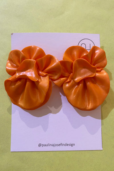Two shiny, orange, flower-shaped Paulina Josefin design earrings from BUTIK KUBIK are displayed on a white card labeled @paulinajosefindesign against a light green background. Unique pieces for your collection.