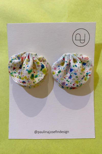 Two round fabric and polymer clay Paulina Josefin design earrings by BUTIK KUBIK, featuring colorful paint splatters, are showcased on a white card with minimalist branding and Instagram handle @paulinajosefindesign against a yellow background.