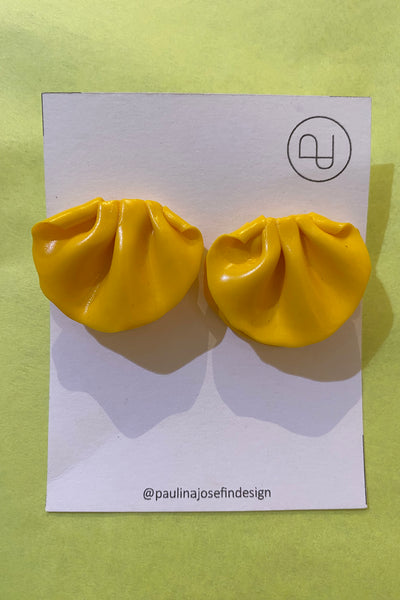 A pair of bright yellow, dumpling-shaped Paulina Josefin design earrings by BUTIK KUBIK, handmade in polymer clay, shown on a white card with logo and @paulinajosefindesign handle, set against a light green background.