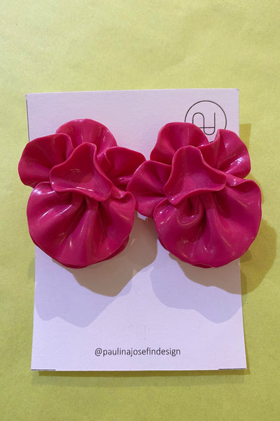 A pair of shiny, bright pink ruffled floral Paulina Josefin design earrings by BUTIK KUBIK is displayed on a white card with the designer's logo and Instagram handle @paulinajosefindesign against a yellow background.