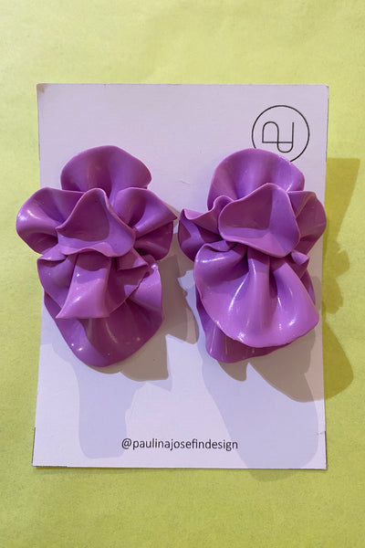 A pair of shiny, ruffled, lavender Paulina Josefin design earrings by BUTIK KUBIK is displayed on a white card with a round logo and @paulinajosefindesign text at the bottom, set against a light yellow background.