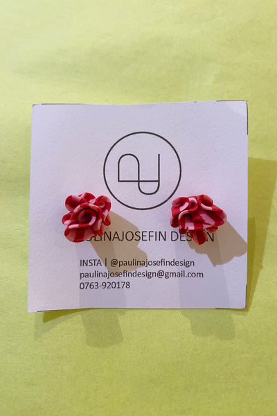A pair of red, flower-shaped Paulina Josefin design earrings by BUTIK KUBIK rest on a white card with logo and contact info, set against a pale yellow background.