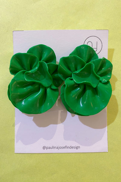 A pair of bright green, ruffled flower-shaped polymer clay Paulina Josefin design earrings by BUTIK KUBIK, displayed on a white card with @paulinajosefindesign at the bottom against a yellow background.