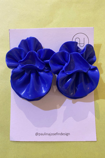 A pair of bright blue, flower-shaped Paulina Josefin design earrings by BUTIK KUBIK with ruffled petals sit on a white card featuring @paulinajosefindesign and logo, set against a light yellow background.