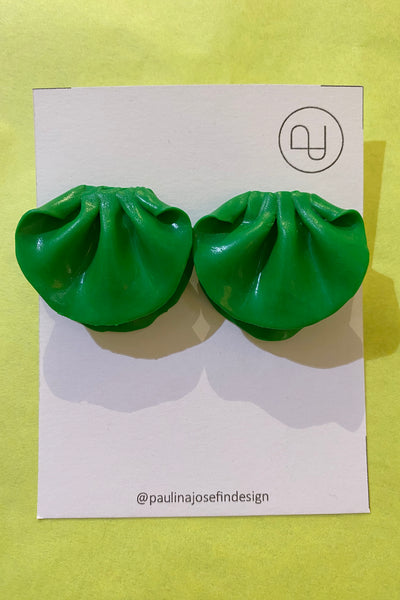 Two green dumpling-shaped Paulina Josefin design earrings by BUTIK KUBIK, made from polymer clay, are shown on a white card with a circular logo and @paulinajosefindesign text, set against a bright yellow background.