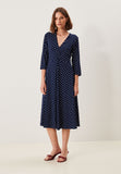 A woman stands against a plain background wearing the Jumperfabriken Dress Noelle in Midnight Blue from BUTIK KUBIK—featuring white polka dots, three-quarter sleeves, and a V-neckline—paired with red sandals.