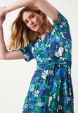 A woman poses with raised arms in the Jumperfabriken Dress Michaela from BUTIK KUBIK, a short-sleeved wrap dress in vibrant blue and green floral print made from organic cotton with white, blue, and green flowers.
